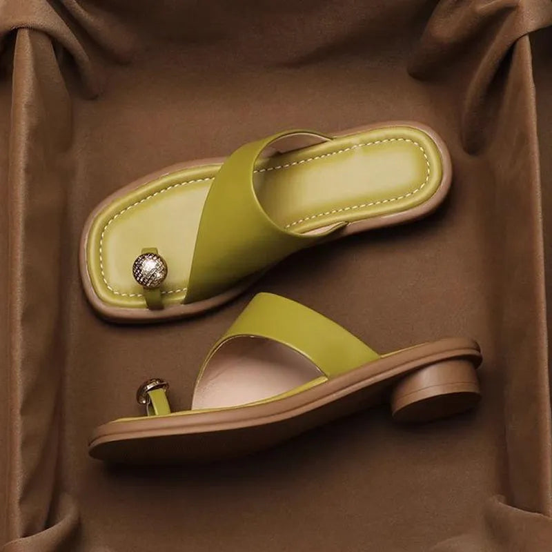 A pair of stylish green Square Toe Slides with a small round metallic embellishment near the big toe, displayed on a brown fabric background. These fashionable slides add a modern twist to any outfit.