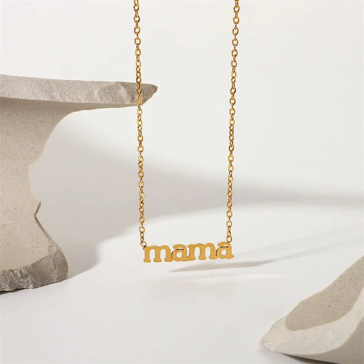 A gold mama pendant necklace with the word mama in lowercase letters hangs between two abstract pieces of light-colored stone on a white background. Perfect as a Mothers Day necklace for celebrating moms every day.