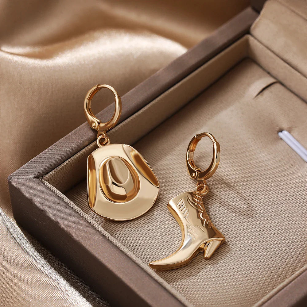 A pair of gold cowboy hat earrings and a boot, perfect as cowgirl accessories, displayed on soft beige satin fabric in a jewelry box.