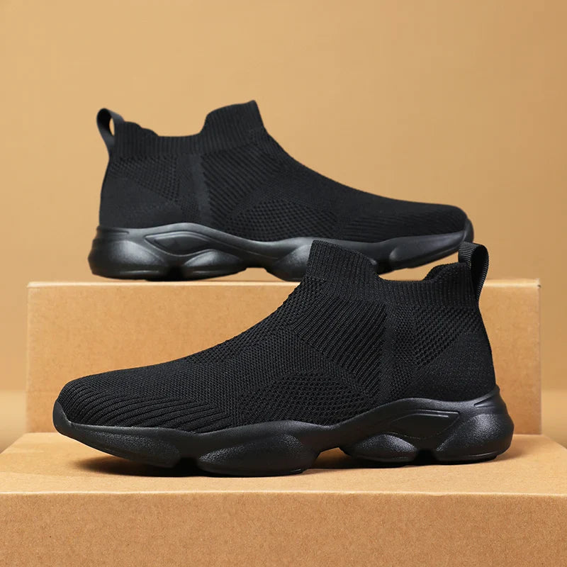 A pair of black slip-on athletic shoes with a breathable knit fabric upper and chunky cushioned, non-slip sole displayed on cardboard boxes against a beige background.