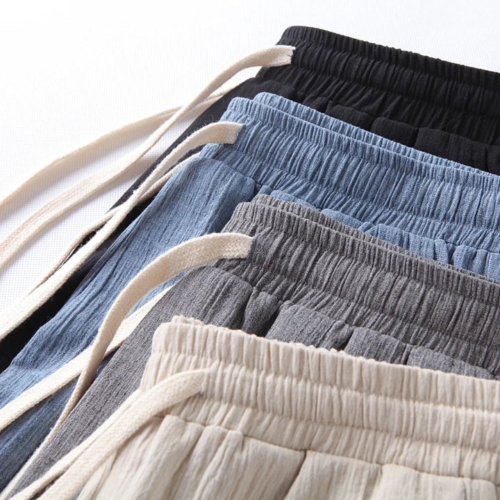 Five pairs of comfortable pants made from pure cotton in beige, gray, blue, dark blue, and black are laid out in a fan pattern, showcasing their elastic waistbands and cotton drawstrings for a modern design look.