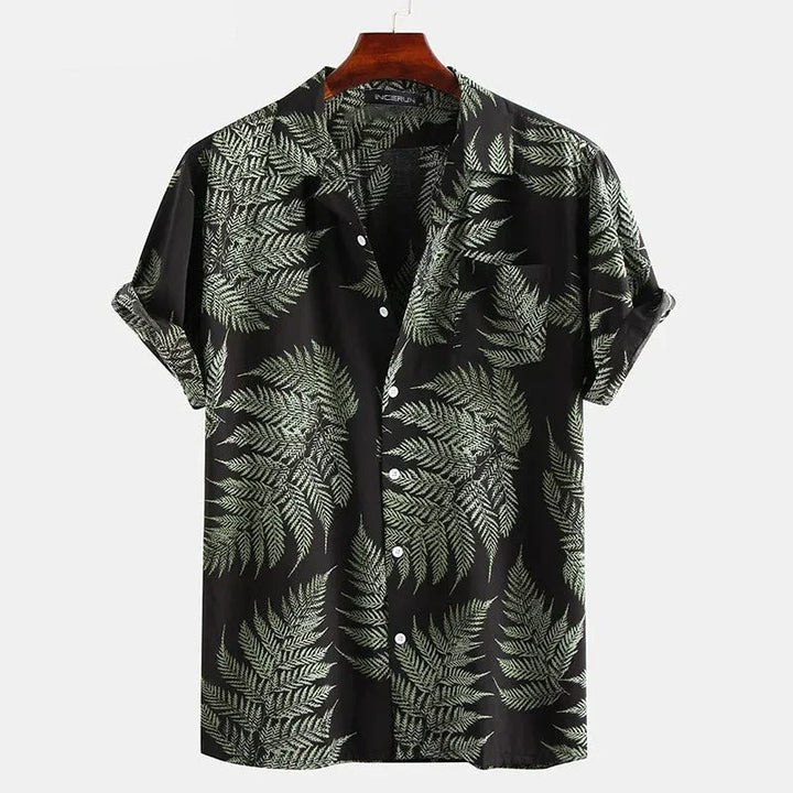A classic design, short-sleeve cotton shirt with a black background and a green fern leaf pattern. Perfect for a casual brunch, its displayed on a wooden hanger against a plain white background.