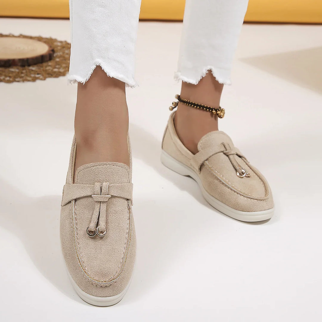 A person wears light beige chic moccasins with tassel details and white frayed-hem pants. An anklet with small charms adorns one ankle. The neutral background features part of a woven mat.