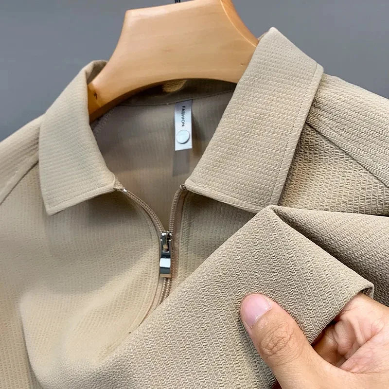 A close-up of a beige textured silk polo shirt on a hanger, with a hand gently pinching the sleeve to highlight the fabric’s moisture-wicking texture.