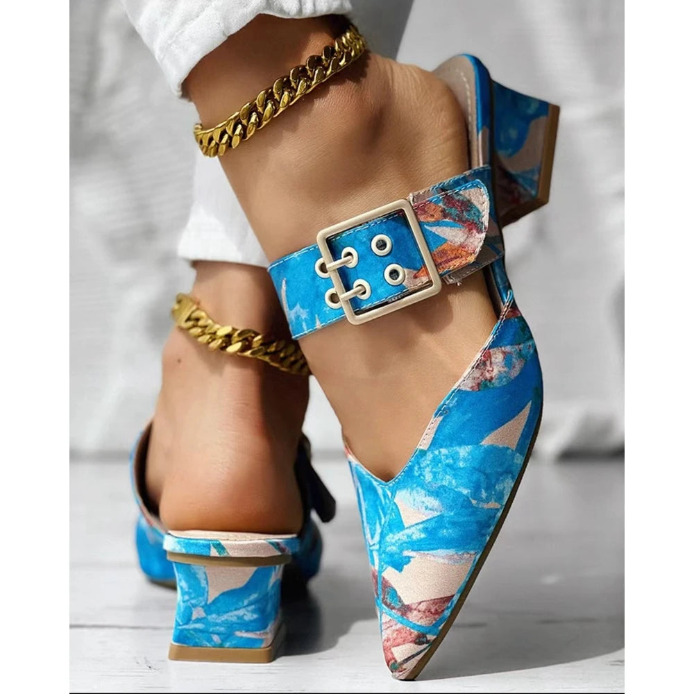 A person wears vibrant blue patterned square heel mules slippers with a large buckle and gold anklets. Paired with white pants, these summer shoes showcase a lively, colorful design perfect for warm days.