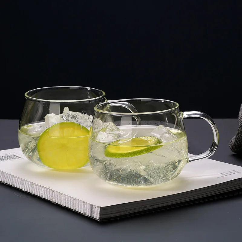 Two clear glass cups with ergonomic handles, filled with ice, water, and lime slices, rest atop a closed white book against a dark background.