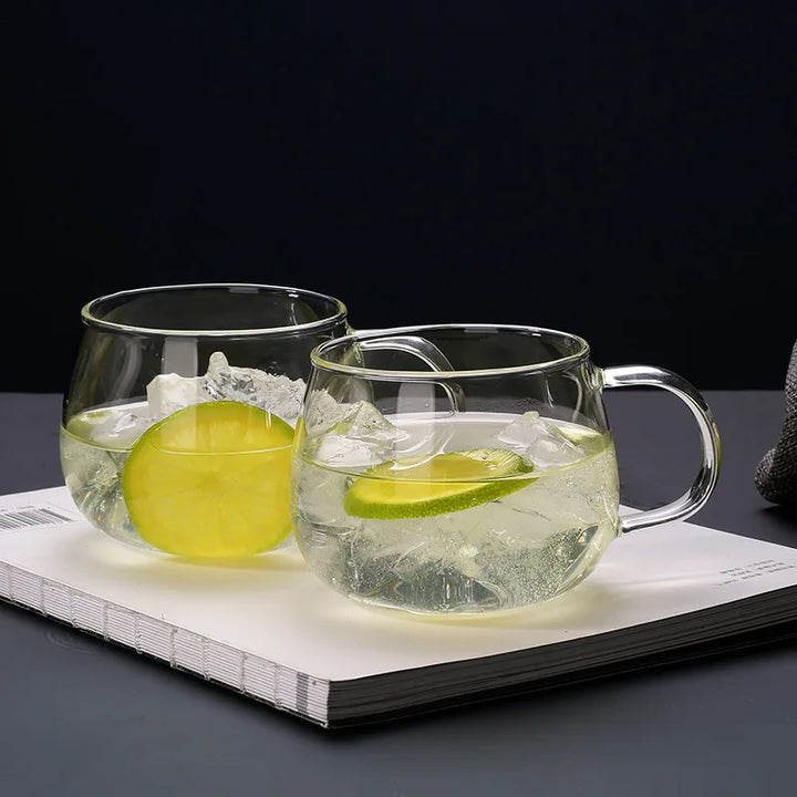Two clear glass cups with ergonomic handles, filled with ice, water, and lime slices, rest atop a closed white book against a dark background.