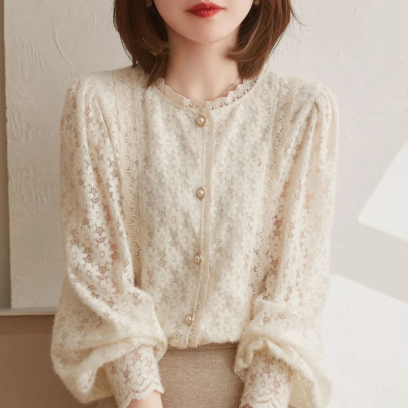 Woman wearing a delicate ivory lace blouse with a floral lace pattern and vintage pearl buttons, featuring slightly puffed long sleeves, sits against a light textured background. Her face is partially visible.