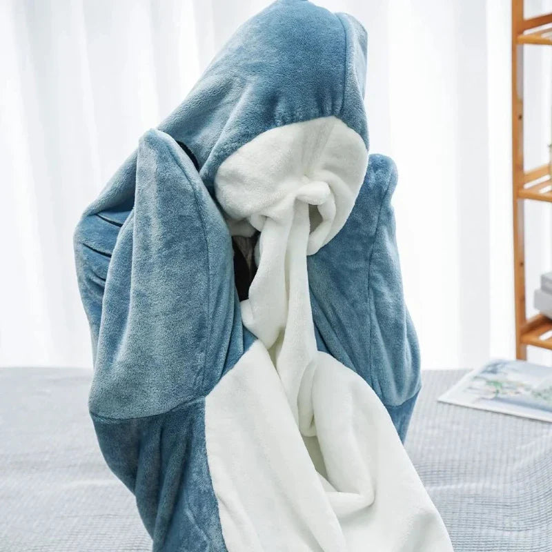 A person is wrapped in a soft, blue and white blanket that resembles shark pajamas, with the blanket covering their head and most of their body. The background shows a table and part of a shelf.