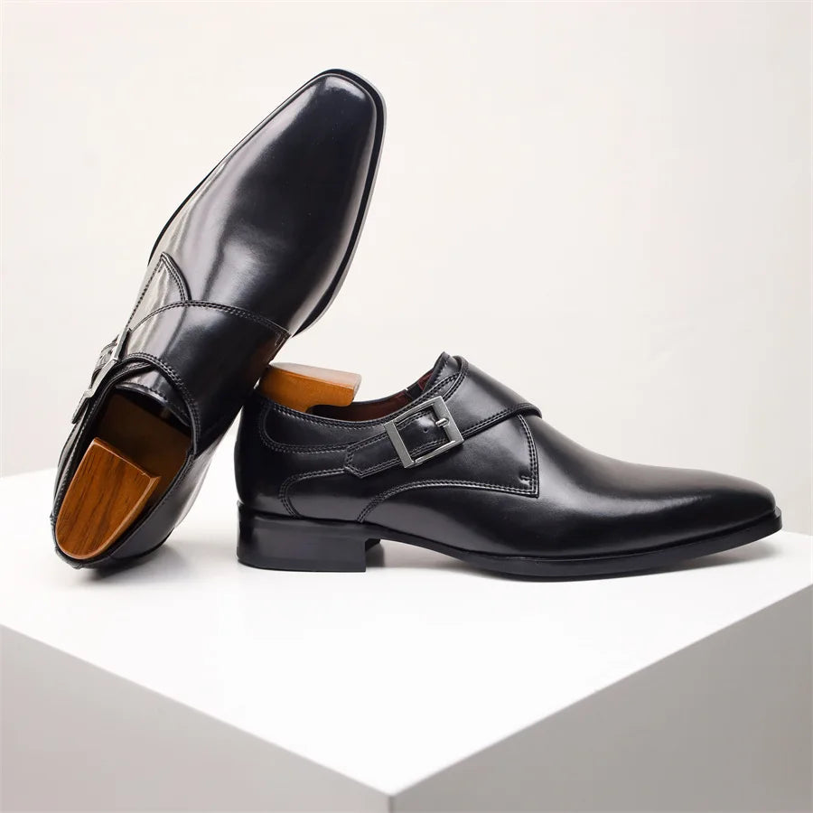 A pair of shiny black Italian leather monk strap shoes with silver buckles, one standing upright and the other leaning against it, displayed on a white cube against a plain background.