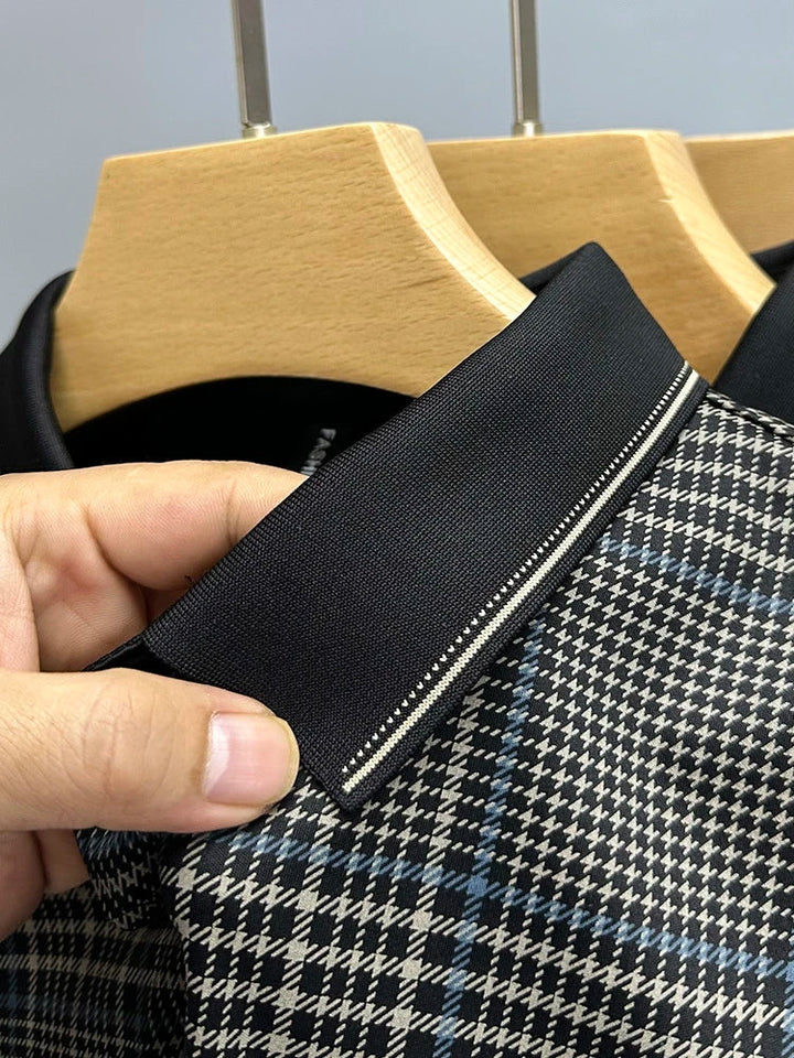 A close-up of a hand holding the collar of a plaid shirt on a wooden hanger. The breathable shirt features a black collar with a thin white stripe and a black, white, and blue houndstooth pattern.