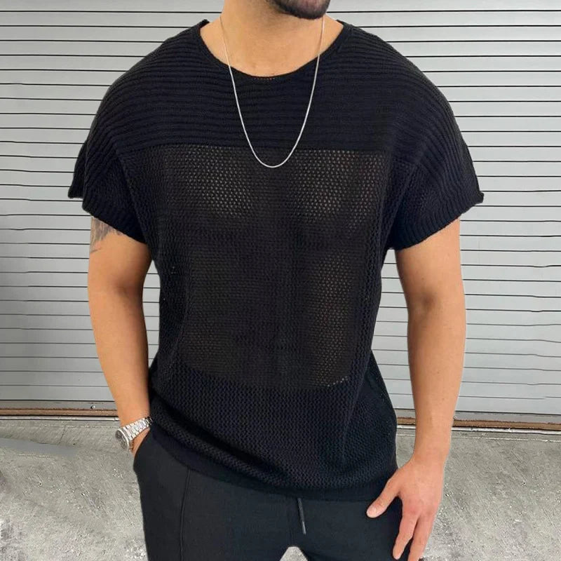 A man wearing a black mesh knit tee, black pants, a silver chain necklace, and a silver watch stands in front of a gray, horizontal-striped wall with his hands in his pockets, showcasing modern streetwear style.