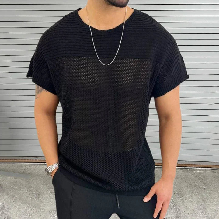 A man wearing a black mesh knit tee, black pants, a silver chain necklace, and a silver watch stands in front of a gray, horizontal-striped wall with his hands in his pockets, showcasing modern streetwear style.