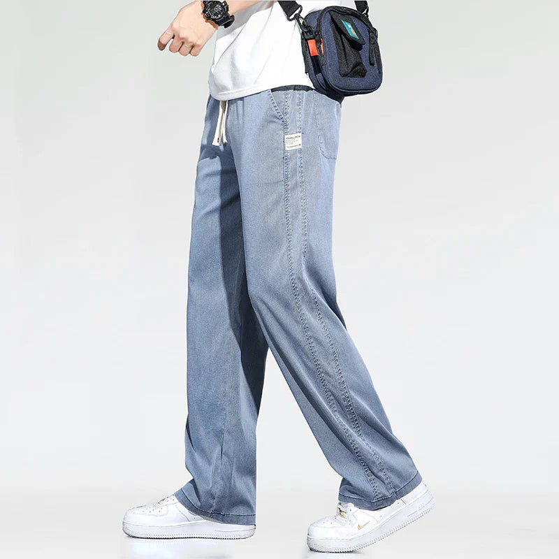 A person wearing light blue Summer Loose Men Jeans, a white t-shirt, white sneakers, a black watch, and carrying a navy blue crossbody bag is walking against a plain light background.