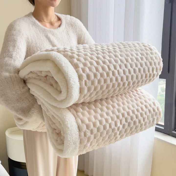 A person wearing a soft, light-colored outfit is holding two large, ultra-soft cotton blankets with a textured, waffle-like pattern, standing near a window with sheer white curtains.