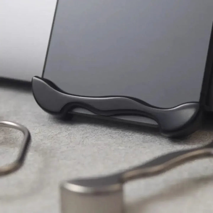 Close-up of a black phone stand holding a smartphone upright on a gray surface, featuring an Aluminum Alloy Case and blurred metal objects in the foreground.