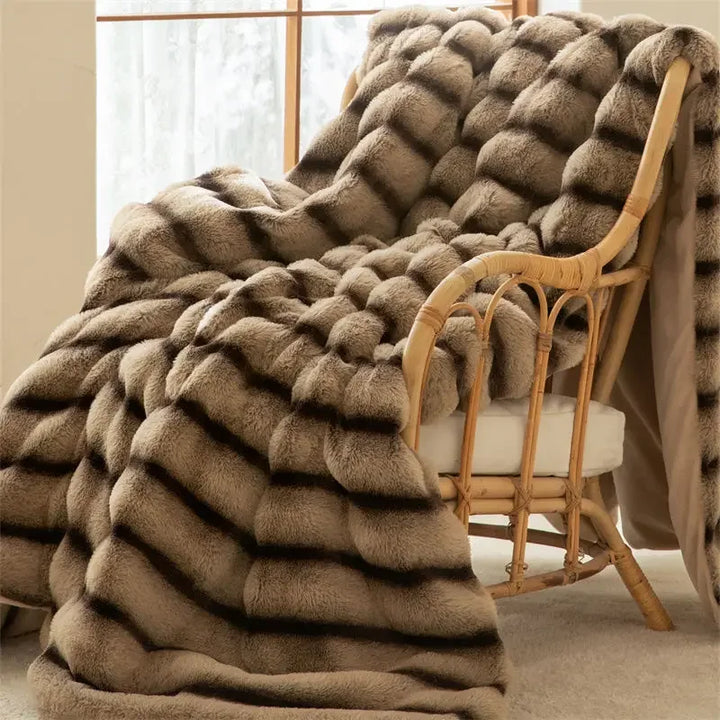 A plush, striped faux rabbit fur blanket with an elegant wave pattern in shades of brown drapes over a cushioned wicker chair near a window, creating a cozy and inviting scene.