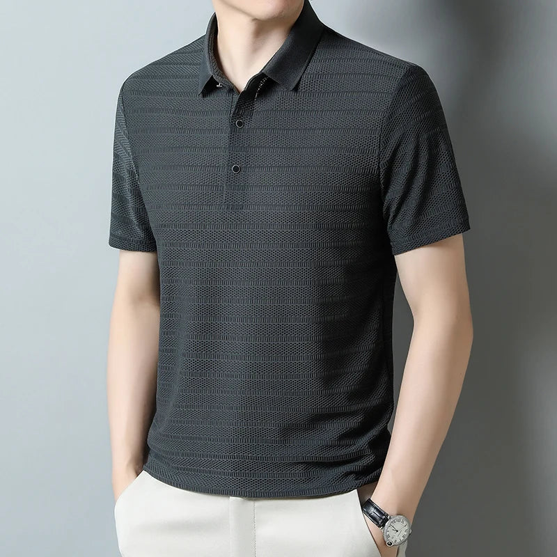 A man wearing a classic polo made from dark, textured silk fabric and light-colored pants stands with his hands in his pockets. He is also wearing a wristwatch and is posed against a plain, light background.