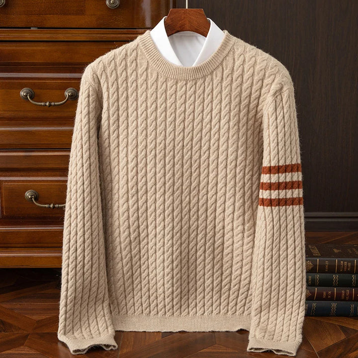 A beige cable-knit sweater with ribbed cuffs, a crew neck, and three brown stripes on the left sleeve, displayed on a hanger over a white shirt, set against a wooden dresser and books.