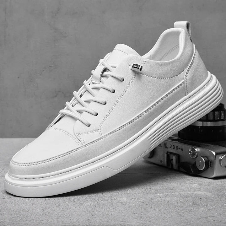 A white sneaker with a thick sole sits on a gray surface, partially resting on a vintage camera. Crafted from genuine leather, the shoe features white laces, a subtle tag on the side, and a minimalist, modern design.