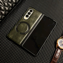 A foldable smartphone in a green Samsung Galaxy Z Fold case with circular MagSafe markings lies partially open on a textured black surface, next to a brown leather-strap wristwatch.