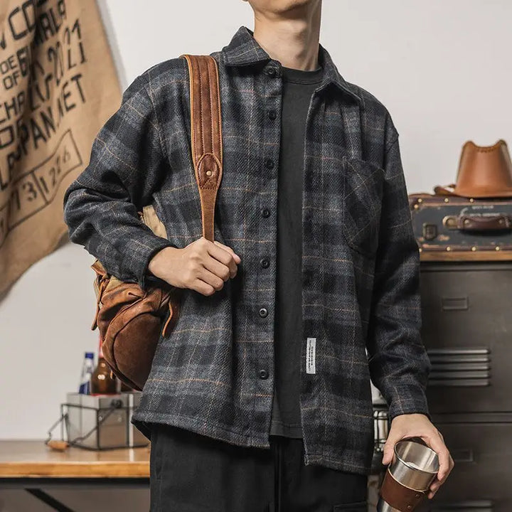 A person wearing an oversized black and gray plaid shirt, holding a metal tumbler in one hand and a brown leather backpack over one shoulder, stands indoors near stacked suitcases and a tan bag hanging on the wall.