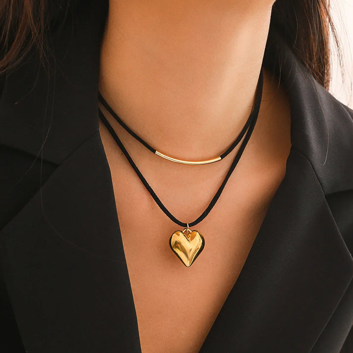 A woman wearing a black blazer and two black cord necklaces: one is an adjustable velvet choker with a gold bar, and the other features a vintage heart pendant.