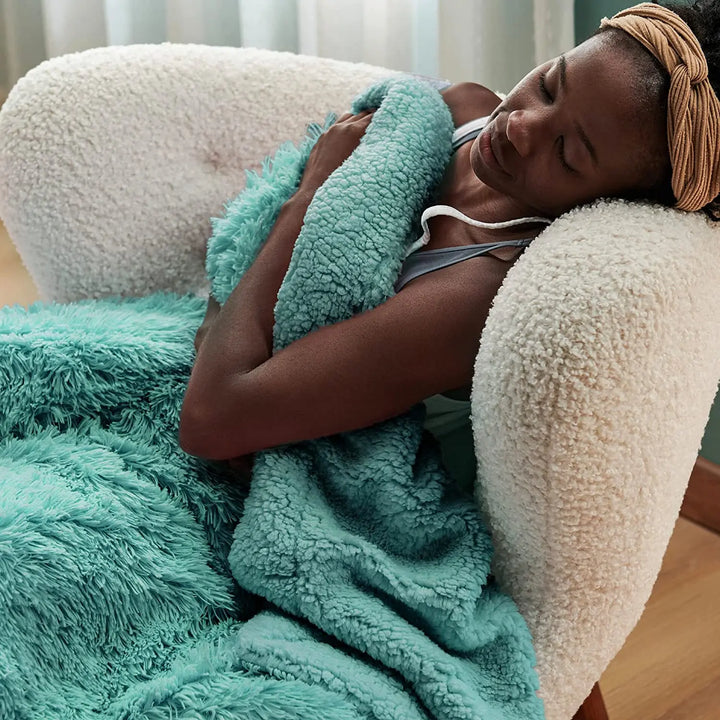 A woman rests in a cozy white armchair, wrapped in a fluffy sage green sherpa blanket. She has her eyes closed and wears a tan headband, appearing calm and comfortable.