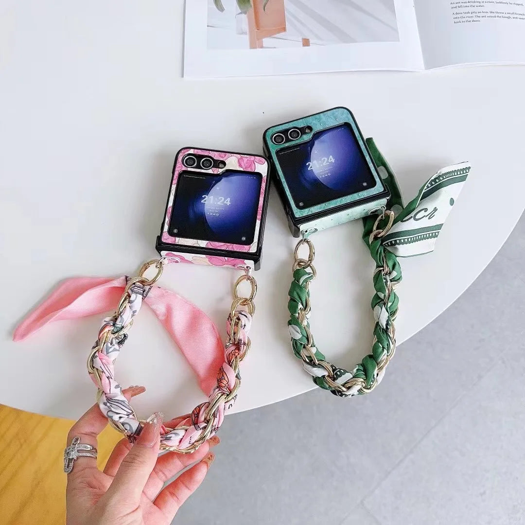 Two foldable smartphones with decorative cases and matching chain straps rest on a white table. One slim design case is pink, the other green. A hand holds the pink case; an open magazine is partially visible nearby.