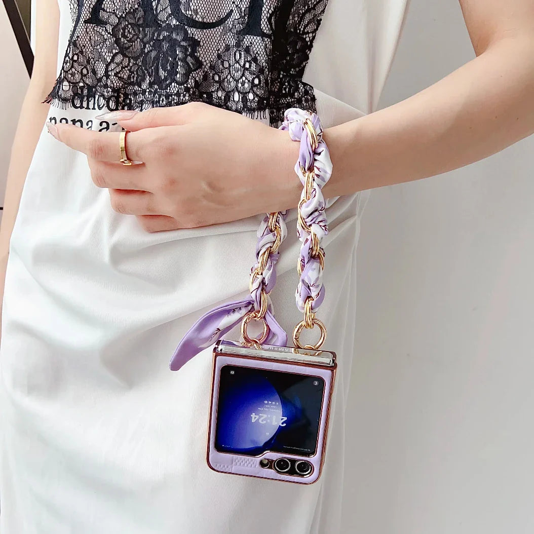 A person wearing a sheer black lace top and white skirt holds a Samsung Galaxy Z Flip in a shiny pink case with a braided lavender wrist strap, offering shockproof protection. The phone screen is visible, and the person wears a gold ring.