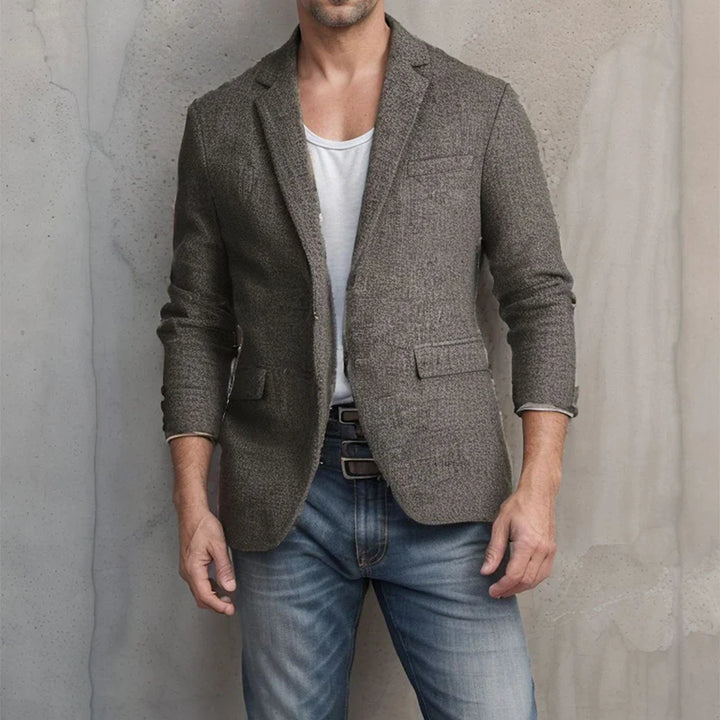 A man showcases timeless style in a gray tweed blazer with a tapered fit over a white T-shirt and blue jeans, standing against a textured, light-colored wall. His head is not visible in the image.