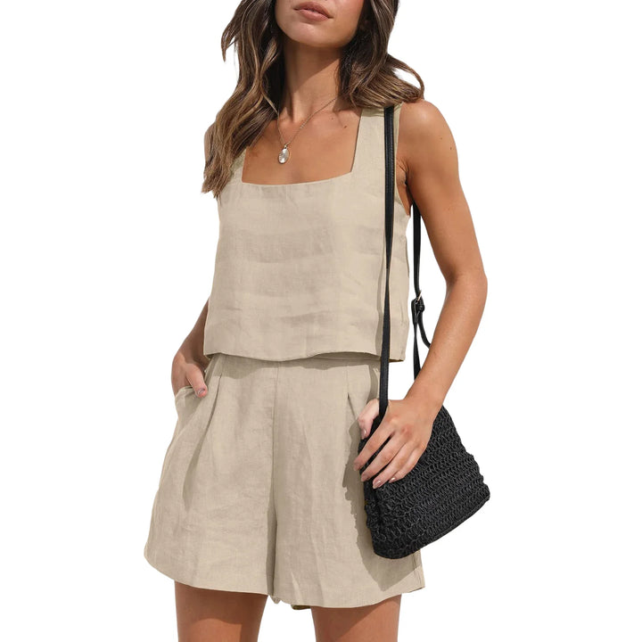 A woman in a sleeveless beige linen set with shorts and a square neckline top, holding a black woven shoulder bag, poses with one hand in her pocket against a white background for a chic contemporary style.
