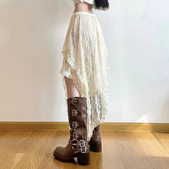 A person wearing a cream-colored, high waist asymmetrical lace skirt and brown knee-high boots with multiple buckles stands on a wooden floor against a plain white wall.