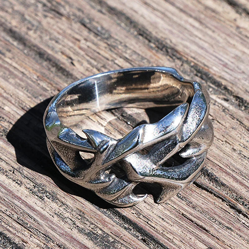 A silver streetwear ring with a bold, interwoven thorn design rests on a weathered wooden surface. The ring features sharp, textured edges, giving it a rugged and intricate fashion jewelry appearance.