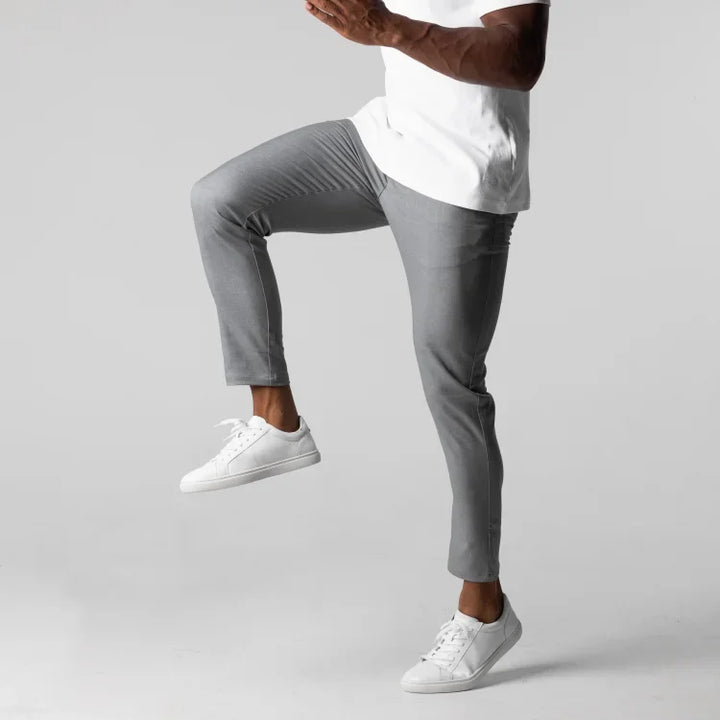 A person wearing gray slim fit, ankle length pants with 4 way stretch, a white shirt, and white sneakers raises one knee in a walking pose against a plain gray background. The person’s face is not visible.