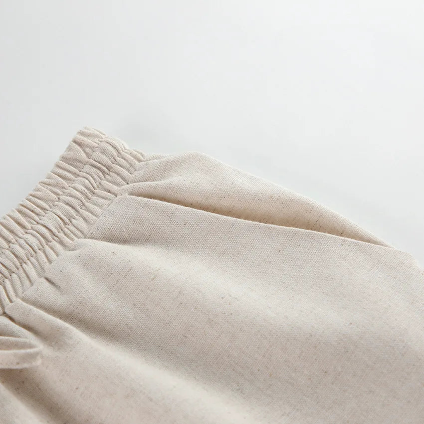 Close-up of the waistband of beige, elastic-waist, wide-leg pants made from a natural, textured fabric, laying flat against a white background.