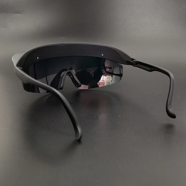 A pair of futuristic, black sunglasses with a single wide, reflective lens and curved arms—offering panoramic visibility—displayed on a dark surface with a shadowy background.