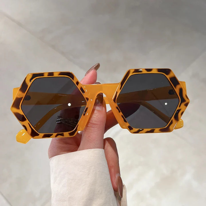 A hand holds yellow, hexagonal frame sunglasses with a tortoiseshell pattern and dark lenses, featuring UV400 protection, against a neutral tiled background.