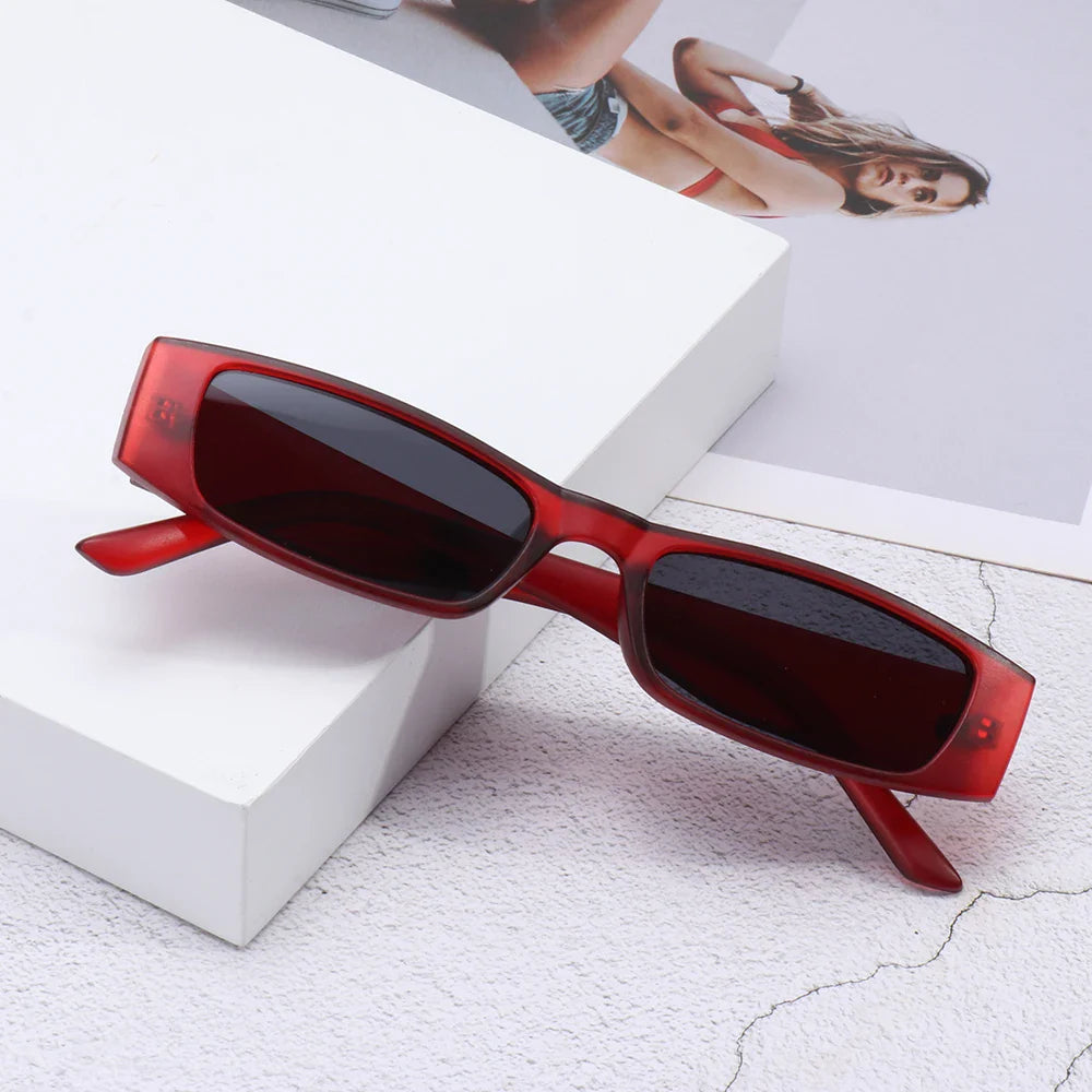 Red retro rectangle sunglasses with dark lenses and a lightweight frame rest on a white surface, with a photo of a woman in swimwear partially visible in the background.