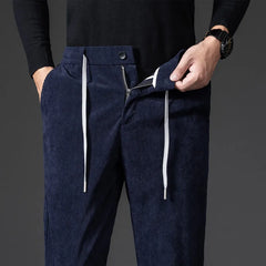 A person wearing a black sweater and watch shows off men's smart casual corduroy pants in navy blue with white drawstrings and a visible zipper fly, one hand casually tucked into the pocket.
