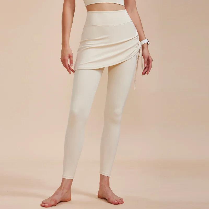 A woman stands barefoot wearing cream-colored high-waisted leggings with an attached, ruched wrap skirt—an effortless addition to any autumn wardrobe. The background is a neutral beige, and her face is not visible in the image.