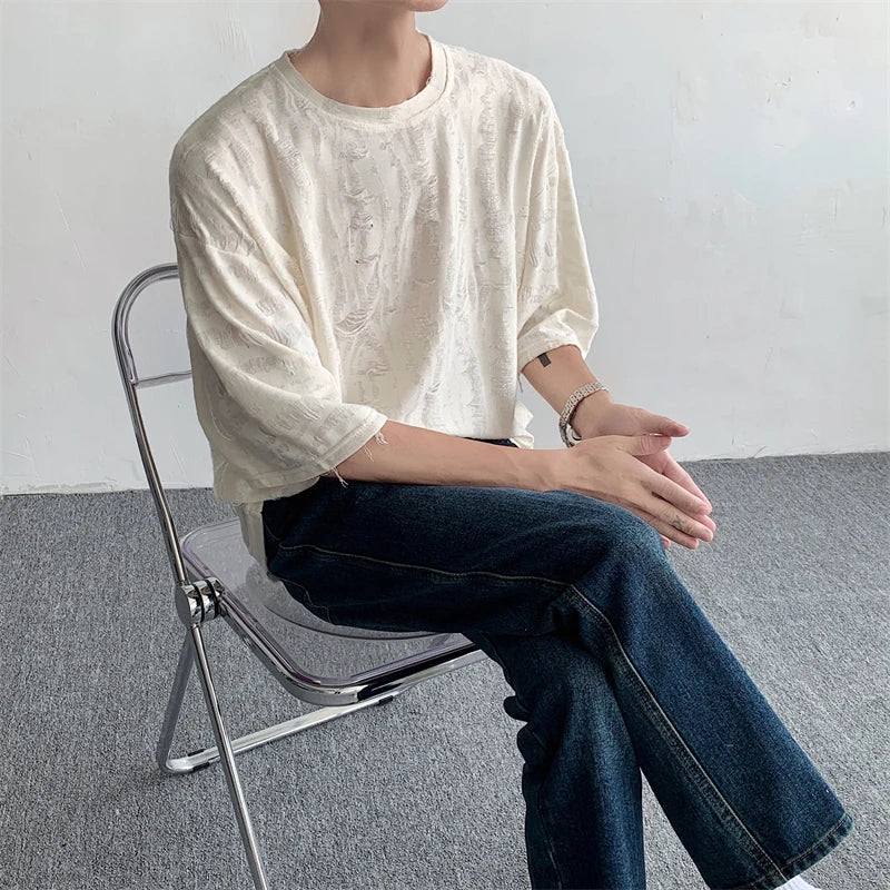 A person wearing an oversized white textured tee and dark jeans sits on a transparent chair with hands clasped, facing sideways against a plain indoor background, channeling effortless streetwear style.