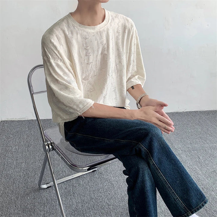 A person wearing an oversized white textured tee and dark jeans sits on a transparent chair with hands clasped, facing sideways against a plain indoor background, channeling effortless streetwear style.