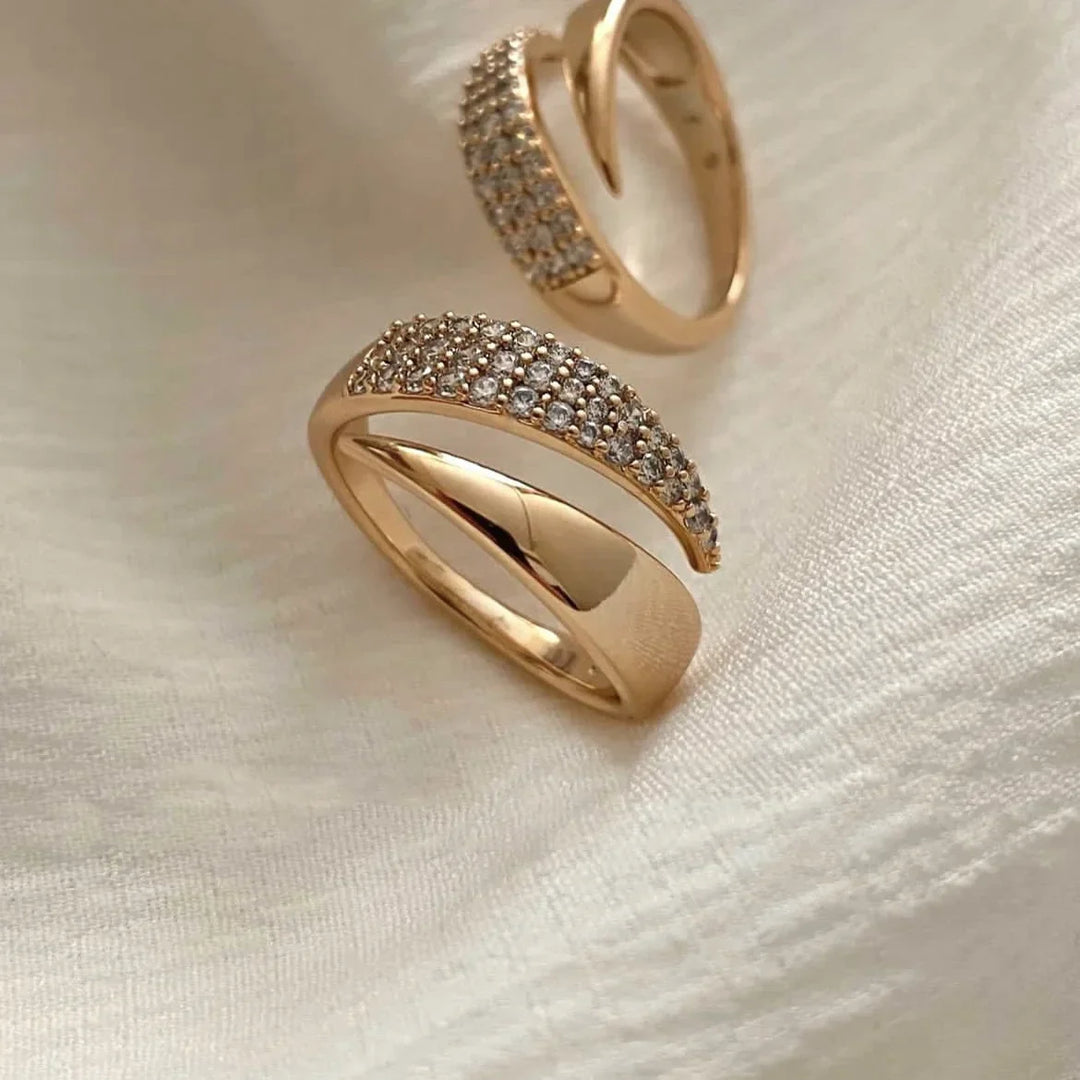 Two gold rings rest on a soft, light-colored fabric. One boasts a polished finish, while the other showcases pavé diamonds set along its curved top. Both feature a modern, twisted design with overlapping bands.