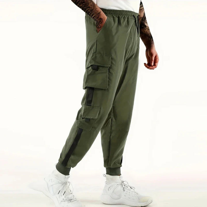A person wearing olive green cargo jogger pants with ribbed ankle cuffs and black accents pairs them with white sneakers, standing against a white background. Only the lower half of their body is visible.