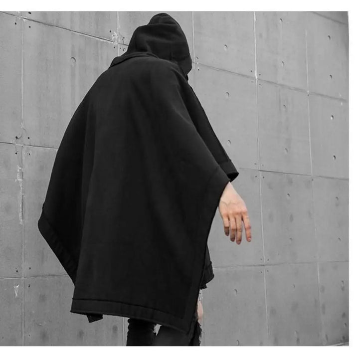 A person wearing a black techwear hoodie stands against a gray concrete wall, with their head turned away and one arm extended downward. The scene has a minimalistic and mysterious atmosphere.