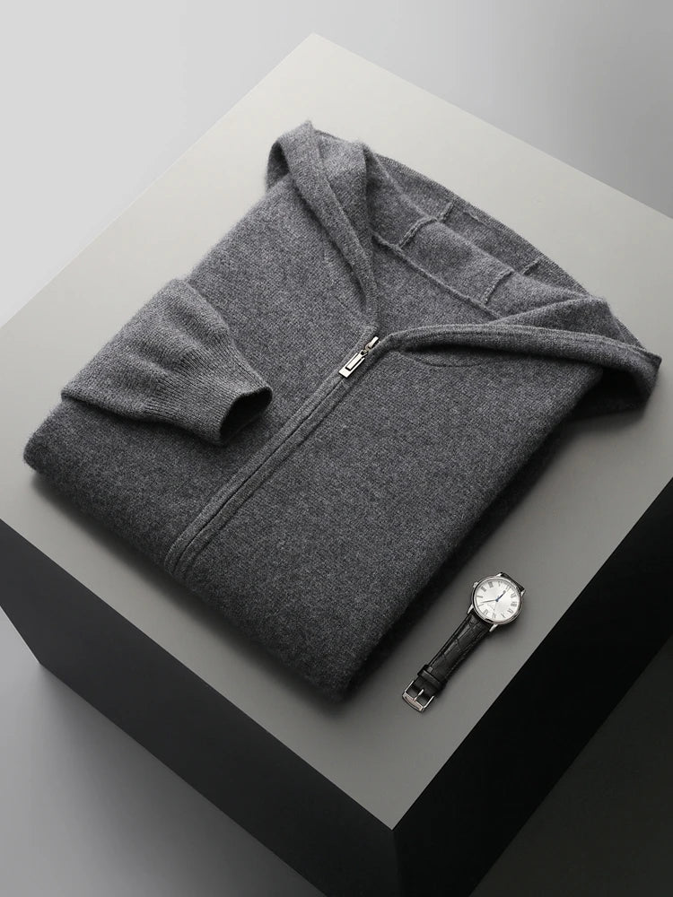 A neatly folded dark gray men wool cardigan with a hood is placed on a light gray platform. A silver wristwatch with a black band lies next to it. The scene is minimal and well-lit.