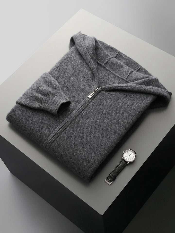 A neatly folded dark gray men wool cardigan with a hood is placed on a light gray platform. A silver wristwatch with a black band lies next to it. The scene is minimal and well-lit.