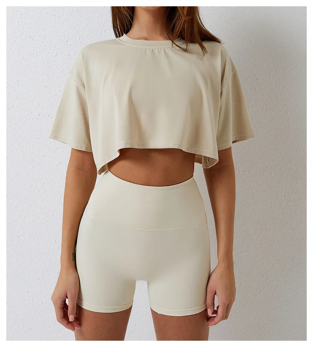 A woman is wearing a cotton crop top and high-waisted, fitted beige shorts, standing against a plain white wall. Her head is partially out of frame.