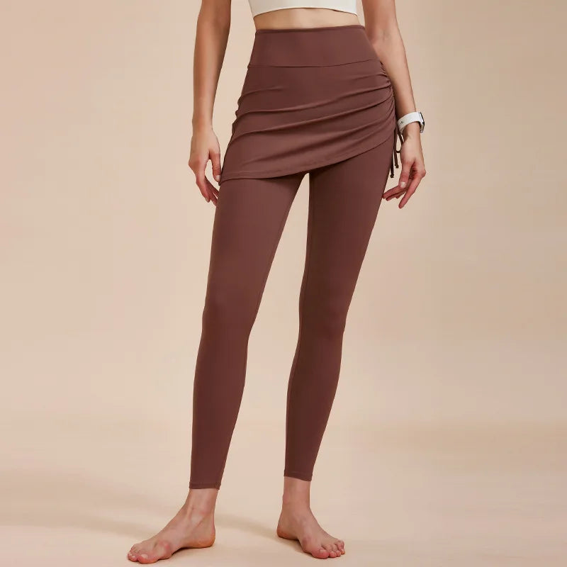 A woman stands barefoot in high-waisted leggings with skirt detailing, paired with a white crop top—an effortlessly chic look for any autumn wardrobe, set against a plain beige background.